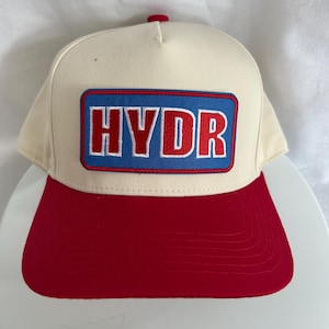 Könnte beinhalten: Cremefarbene Baseballkappe mit rotem Schirm und rotem Knopf oben. Die Kappe hat einen rechteckigen Aufnäher mit dem Wort "HYDR" in roten Blockbuchstaben auf blauem Hintergrund.