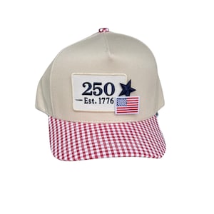 May include: A beige baseball cap with a red and white gingham peak. The cap has a patch displaying the number "250", a blue star, the text "- Est. 1776", and a small American flag.