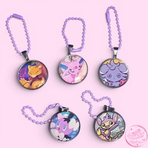 Può includere: Cinque ciondoli rotondi con disegni di personaggi dei cartoni animati, ciascuno con una cornice in metallo argentato e una catena viola. I personaggi sono in vari colori, tra cui giallo, rosa e viola, su sfondo rosa.