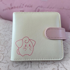 Pode incluir: Uma carteira quadrada bege clara com fecho de pressão rosa. Uma personagem de desenho animado rosa é impressa na frente. A carteira é provavelmente feita de couro sintético e tem uma borda costurada.
