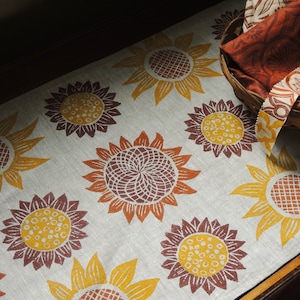 Puede incluir: Un camino de mesa de lino con un patr&oacute;n repetitivo de girasoles marrones y amarillos. El camino de mesa est&aacute; sobre una superficie de madera.