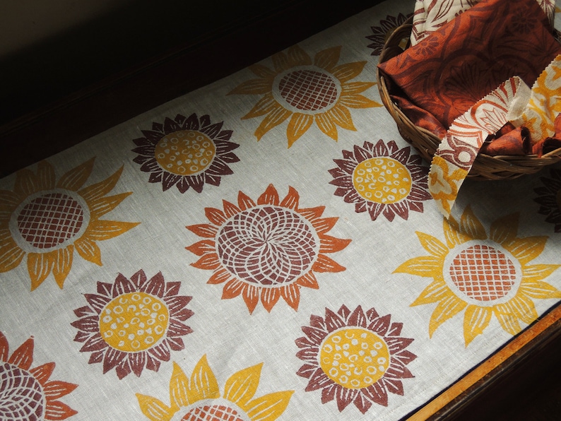 Puede incluir: Un camino de mesa de lino con un patr&oacute;n repetitivo de girasoles marrones y amarillos. El camino de mesa est&aacute; sobre una superficie de madera.