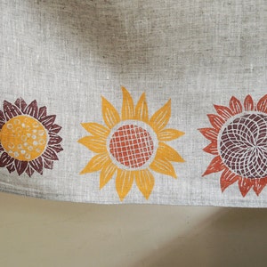 May include: A linen tea towel with a border of four printed sunflowers. The sunflowers are in shades of orange, yellow, brown, and red.