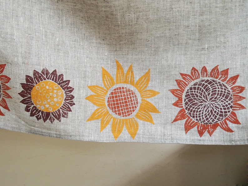 May include: A linen tea towel with a border of four printed sunflowers. The sunflowers are in shades of orange, yellow, brown, and red.