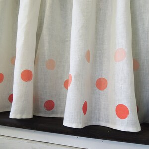 May include: White linen curtains with large, coral-colored polka dots. The curtains are hanging in front of a window, with the fabric slightly gathered. The polka dots vary in size and shade, creating a playful and cheerful design.