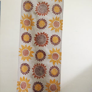 Puede incluir: Un camino de mesa de lino largo con un patr&oacute;n repetitivo de girasoles estilizados en tonos naranja, amarillo y marr&oacute;n.