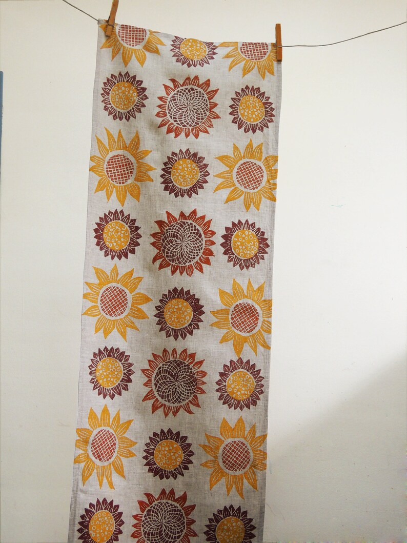 Puede incluir: Un camino de mesa de lino largo con un patr&oacute;n repetitivo de girasoles estilizados en tonos naranja, amarillo y marr&oacute;n.