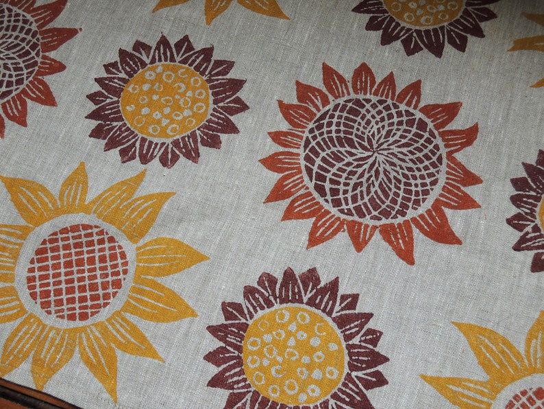 Puede incluir: Primer plano de una tela con un patr&oacute;n repetitivo de girasoles estilizados en tonos naranja, amarillo, marr&oacute;n y rojo. La tela tiene una textura similar al lino.