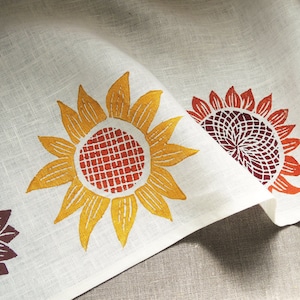 May include: White linen fabric with a repeating pattern of orange, yellow, and red sunflowers. The fabric is folded in half, showing the pattern on both sides.