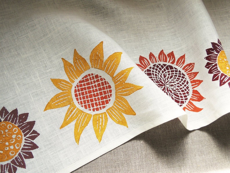 May include: White linen fabric with a repeating pattern of orange, yellow, and red sunflowers. The fabric is folded in half, showing the pattern on both sides.