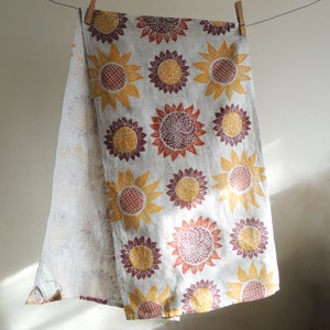 Puede incluir: Un pa&ntilde;o de cocina de lino con un estampado de girasol en tonos amarillos, naranjas y marrones. El pa&ntilde;o tiene un borde con flecos y est&aacute; colgado de dos pinzas de madera.