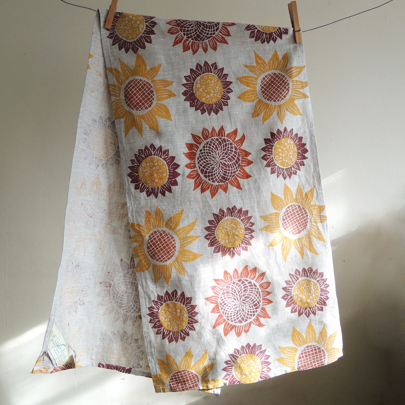 Puede incluir: Un pa&ntilde;o de cocina de lino con un estampado de girasol en tonos amarillos, naranjas y marrones. El pa&ntilde;o tiene un borde con flecos y est&aacute; colgado de dos pinzas de madera.