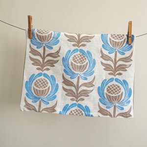 May include: A white linen tea towel with a blue and brown floral pattern. The towel is hanging on a clothesline with wooden clothespins.