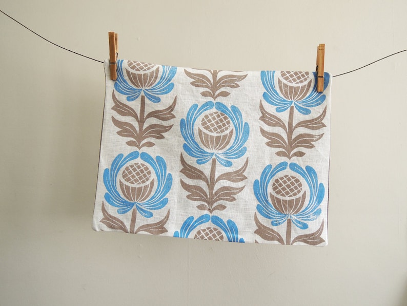 May include: A white linen tea towel with a blue and brown floral pattern. The towel is hanging on a clothesline with wooden clothespins.