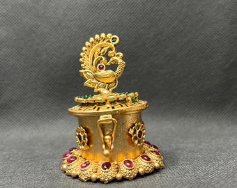 May include: A gold-colored metal box with a peacock design on the lid. The box is decorated with red and green gemstones.