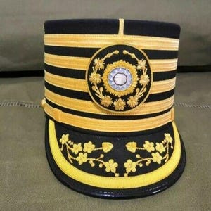 Op de afbeelding: Een zwarte en gouden militaire hoed met een gouden geborduurd bloemenpatroon en een zilveren knoop.