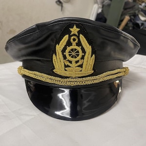 Op de afbeelding: Een zwarte leren pet met een gouden galonversiering en een geborduurd embleem in goud met een wiel en bladeren.