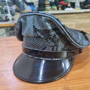 Könnte beinhalten: Eine schwarze und graue Lederkappe mit Schlangenhautmuster. Die Kappe hat ein schwarzes Lederband mit glänzender Oberfläche.