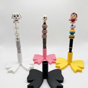 May include: Three decorative pen holders with bow-shaped bases in white, pink, yellow, and black. Each holder features a different character or design on top, including a Dalmatian puppy, a bunny, and a cartoon character. The pens are designed to stand upright.