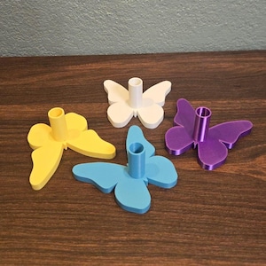May include: Four butterfly-shaped candle holders in yellow, white, blue, and purple. Each holder has a cylindrical center for a candle. The holders are made of a smooth material and sit on a wooden surface.