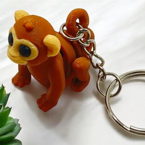 May include: A brown monkey keychain with a silver key ring. The monkey is sitting with its tail curled around its body.