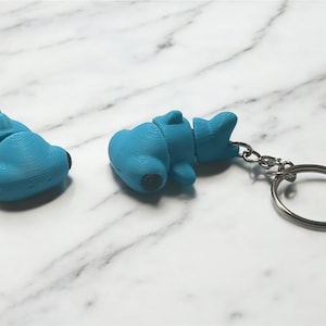 May include: Two blue 3D-printed shark keychains, one with a keyring attached. The shark keychains have black eyes and are lying on a white and gray marble surface. The keychains are a fun accessory.