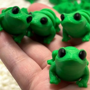 Op de afbeelding: Verschillende kleine, felgroene kikkerbeeldjes met zwarte ogen worden getoond. De kikkers zijn 3D-geprint en hebben een gladde textuur. De kikkers staan in verschillende poses, sommige naar voren en andere opzij.