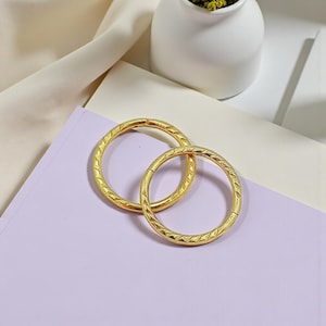 May include: Two gold-coloured metal rings with a textured, repeating pattern. The rings are circular and appear to be made of a shiny, metallic material. They are resting on a light purple surface, with a white vase and a beige fabric in the background.
