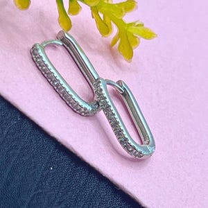 May include: A pair of silver-coloured, rectangular hoop earrings. The outer edges of the hoops are lined with small, clear gemstones. The earrings are displayed on a pink surface with a sprig of yellow flowers.