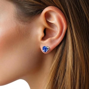 Puede incluir: Primer plano de la oreja de una mujer con un pendiente de botón azul en forma de corazón. El pendiente tiene un engaste plateado y una piedra vibrante de color zafiro. La mujer tiene el pelo largo y castaño claro.