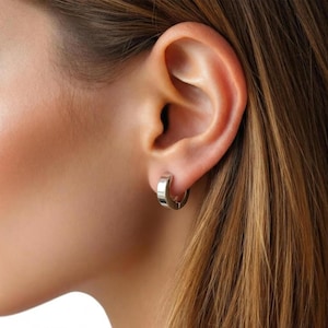May include: A silver hoop earring is shown in close-up, worn on an ear. The earring is a small, thick band with a smooth, reflective surface. The ear is visible, with the earring securely fastened. The background is blurred, focusing attention on the jewelry.