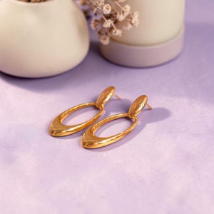 May include: A pair of gold-colored oval drop earrings. The earrings have a polished finish and a post-back closure. The earrings are displayed on a light purple surface, with a blurred background of white vases and dried flowers.