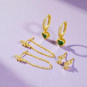 May include: A collection of gold-colored earrings on a purple surface. The earrings include hoop earrings with green heart-shaped pendants, chain earrings, and stud earrings with rectangular stones. The earrings are arranged in a visually appealing display.