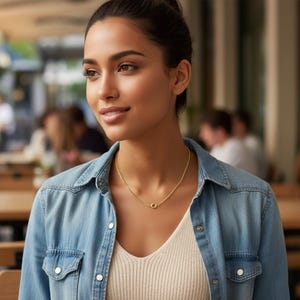 May include: A gold necklace with a small, circular pendant. The person is wearing a light-coloured top and a denim shirt. The background is blurred, suggesting an outdoor setting. The necklace is the focal point.