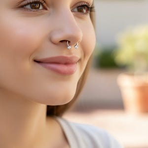 May include: A close-up of a person's face featuring a silver nose ring. The nose ring has two small, clear gemstones. The person is smiling, with a blurred background.