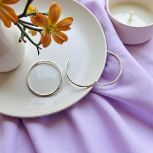 Könnte beinhalten: Zwei silberne Creolen liegen auf einem weißen Teller, neben einer kleinen weißen Vase mit orangefarbenen Blumen. Eine brennende Kerze befindet sich in einer kleinen weißen Tasse. Der Hintergrund ist ein weicher, lavendelfarbener Stoff.