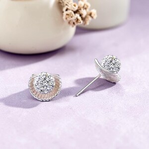 May include: A pair of silver stud earrings with a floral design. Each earring features a cluster of small, sparkling clear crystals at the centre, surrounded by a silver petal-like frame. The earrings are displayed on a light purple surface.
