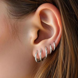 May include: A close-up of an ear adorned with several small, silver hoop earrings. The earrings are evenly spaced and of varying sizes. The ear is set against a backdrop of brown hair.