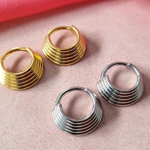 May include: Four gold and silver hoop earrings with a layered design. The earrings are stacked on a pink surface.