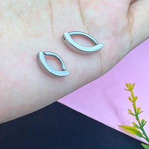 May include: Two silver-coloured, oval-shaped belly button rings. The rings have a curved design with a small clasp. The background includes a hand, a pink surface, and a sprig of greenery.