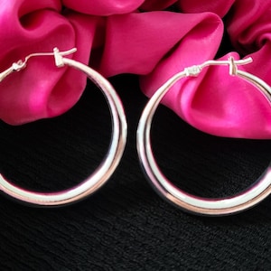Peut inclure: Une paire de boucles d'oreilles créoles en argent avec une finition lisse et arrondie. Les boucles d'oreilles sont présentées sur un fond en tissu rose.