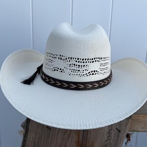 May include: A white cowboy hat with a brown leather band and a black tassel. The hat has a woven design on the crown.
