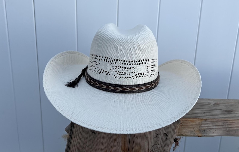 May include: A white cowboy hat with a brown leather band and a black tassel. The hat has a woven design on the crown.