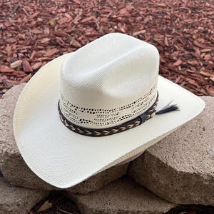 May include: A white straw cowboy hat with a brown leather band and a braided trim. The hat has a decorative pattern around the crown.