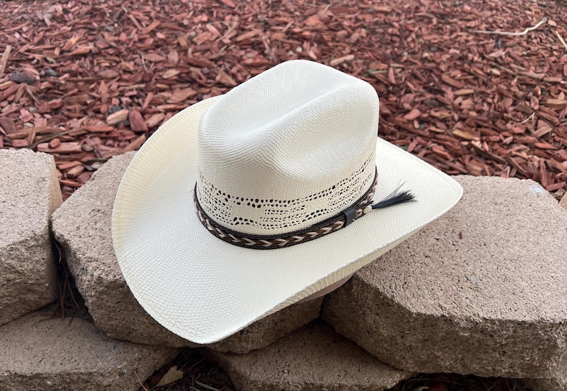 May include: A white straw cowboy hat with a brown leather band and a braided trim. The hat has a decorative pattern around the crown.