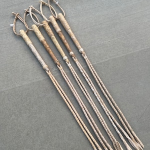 May include: A set of six braided leather whips with a light brown color. The whips are laid out on a gray surface.