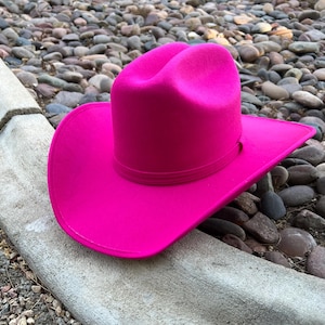 Puede incluir: Un sombrero de vaquero fucsia vibrante con ala ancha y copa estructurada. El sombrero presenta una banda alrededor de la base de la copa. Se encuentra sobre un fondo de piedras lisas y una superficie de hormig&oacute;n.