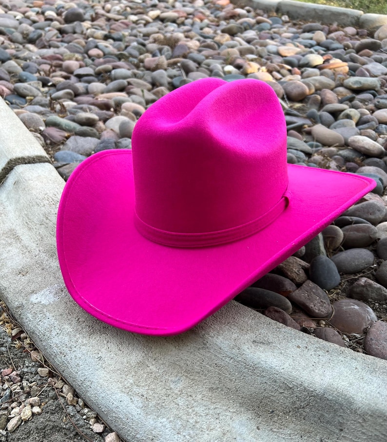 Puede incluir: Un sombrero de vaquero fucsia vibrante con ala ancha y copa estructurada. El sombrero presenta una banda alrededor de la base de la copa. Se encuentra sobre un fondo de piedras lisas y una superficie de hormig&oacute;n.