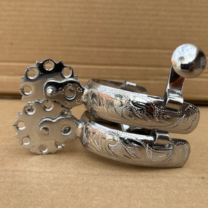 May include: Silver spur with a floral design and a row of holes around the edge. The spur has a rounded tip and a metal band.