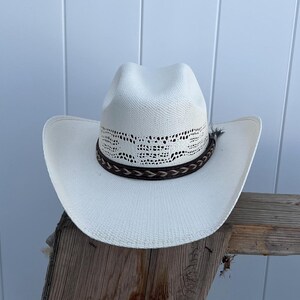 May include: A white straw cowboy hat with a brown leather band and a white lace design. The hat is sitting on a wooden surface.